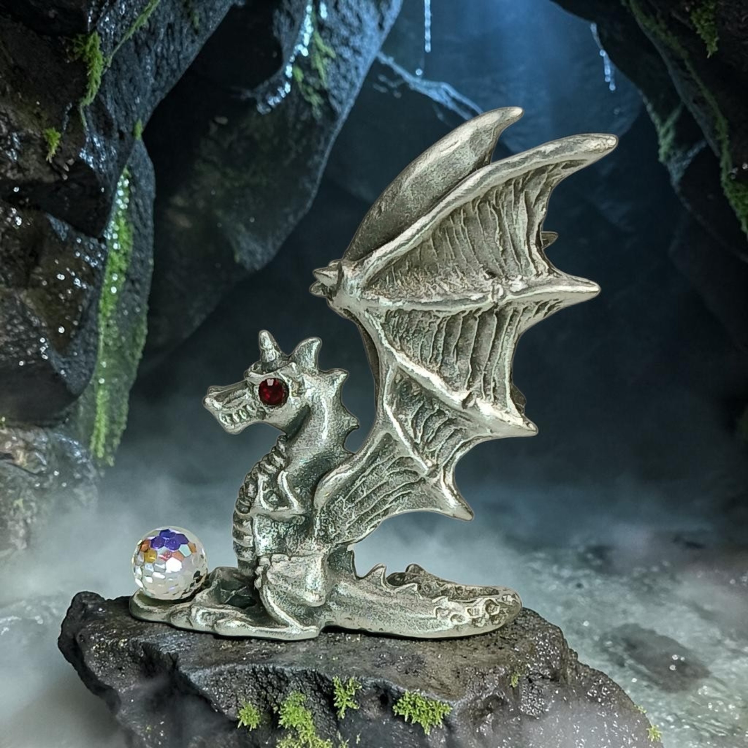 Winged dragon sculpture with a red eye and a curled tail, beside a faceted orb in a cave setting.