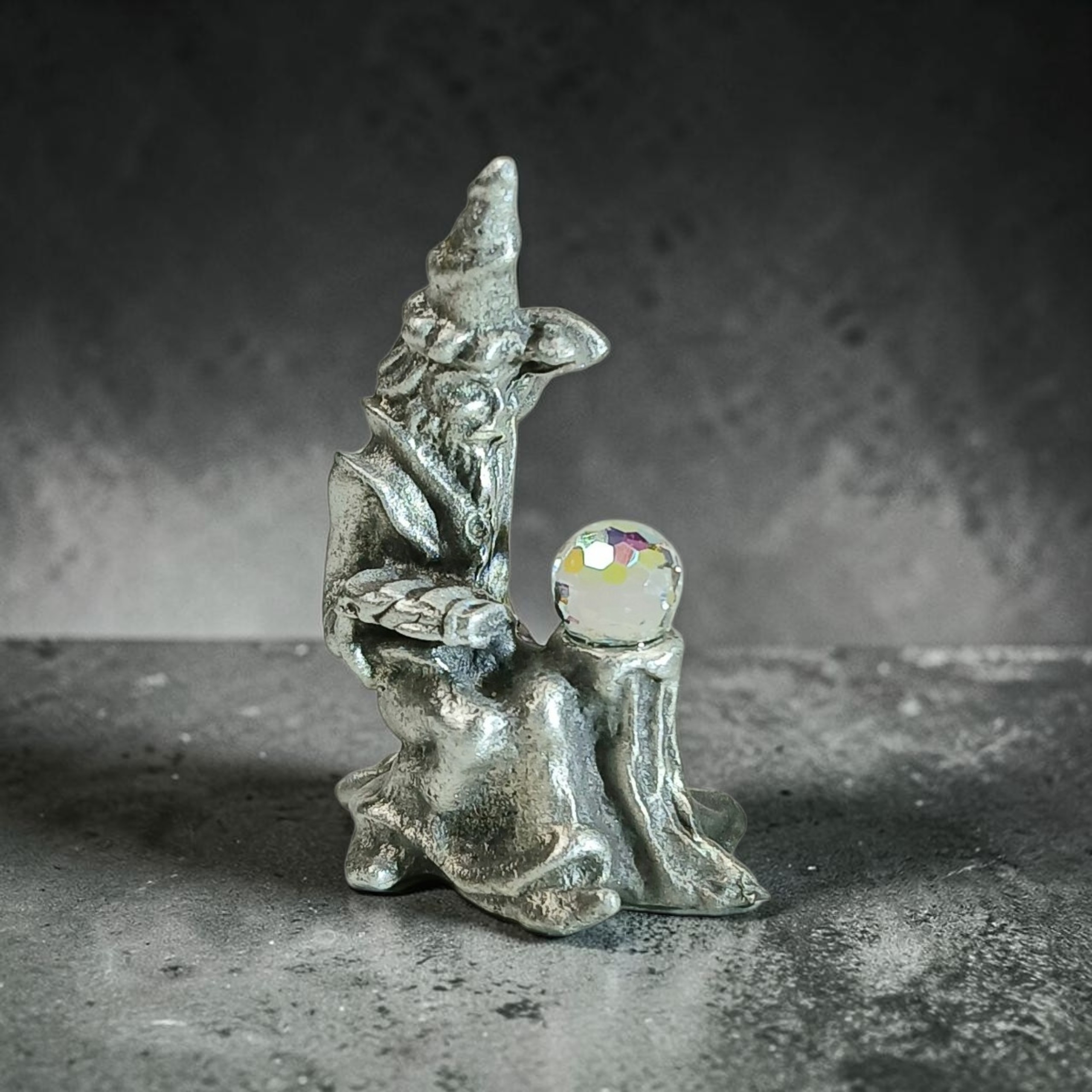 Seated wizard sculpture with a pointed hat and long beard beside a faceted orb on a pedestal.